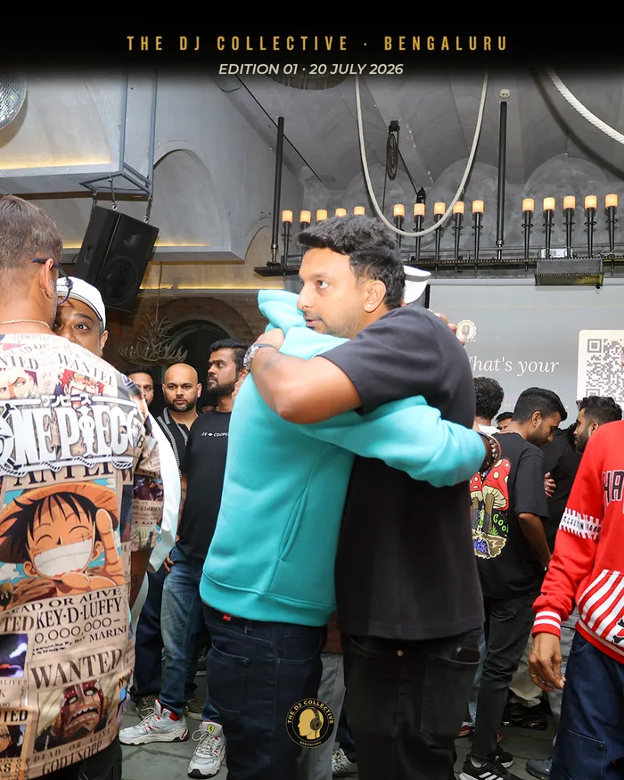 Two DJs mid-hug, one in a teal hoodie, the room busy behind them